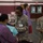 Two U.S. Army soldiers in ACU (Army Combat Uniform) are providing medical assistance at what appears to be a community health event or medical outreach program held in a gymnasium or large indoor facility. One soldier, wearing a stethoscope, is examining a seated civilian patient while a second soldier observes. Additional medical personnel in scrubs are visible in the background.
