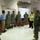 A group of military personnel and civilians are gathered in what appears to be a school auditorium or multipurpose room being used as an emergency operations or command center. A civilian in a yellow safety vest and cowboy hat appears to be briefing the group, gesturing toward a projected screen displaying data. The setting suggests a disaster response or emergency management coordination meeting.