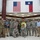 A group of approximately 11 individuals poses for a formal group photo inside what appears to be a school or government building lobby. The group includes military personnel in various uniforms, civilians with ID badges, and one individual in a safety vest and cowboy hat. Two flags — the United States flag and the Texas state flag — are prominently displayed on the wall behind them.