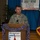 A military officer wearing Texas State Guard uniform with the name tag 'Betty' is speaking at a podium during Operation Lone Star VIP Day 2016. The event appears to be a formal military ceremony or briefing, sponsored by Double Y Orchard. The setting includes patriotic decorations including American and Texas state flags.