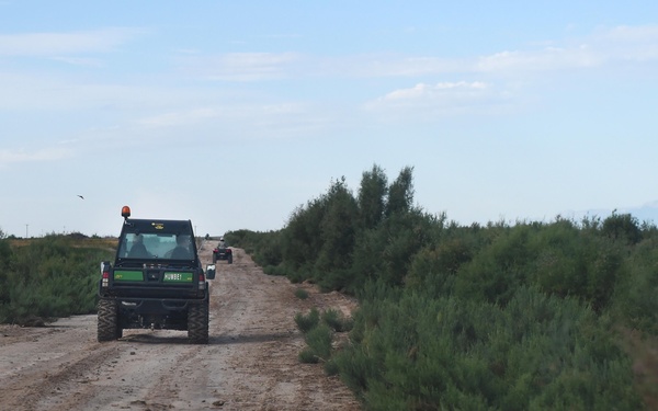 Two off-road utility vehicles are traveling along a narrow dirt track through scrubland or semi-arid terrain. The lead vehicle is a black UTV/side-by-side with green markings, visible from the rear, followed by a second vehicle further down the trail. The landscape is flat with low green shrubs on both sides under a partly cloudy sky.