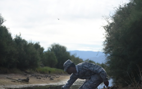 A military service member in digital camouflage uniform is crouched at the edge of a body of water, using a long stick or rod to probe or interact with the water's surface. The scene is set along a muddy riverbank or shoreline with trees and mountains visible in the background. The still water creates a clear reflection of both the soldier and the overcast sky.