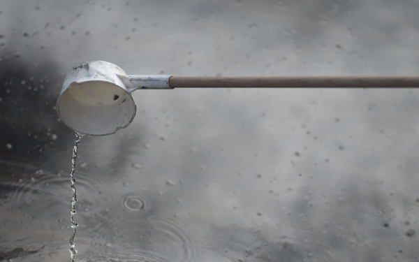 A long-handled ladle or dipper, white in color and mounted on a wooden handle, is pouring water into a basin or pool below. The water stream creates concentric ripple rings on the surface, which appears dark and reflective. This appears to be a traditional Japanese temizuya (hand-washing basin) found at Shinto shrines or Buddhist temples.