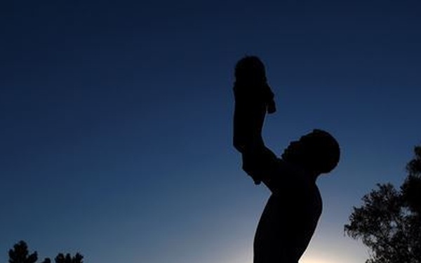 A silhouetted figure stands against a twilight sky, holding a small child or infant raised high overhead with both arms extended upward. The scene is backlit by a setting or rising sun positioned low on the horizon, creating a dramatic contrast between the dark foreground and the gradient blue sky. Trees and rooftop structures are visible in the background as dark silhouettes.