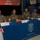 Four military personnel in camouflage uniforms are seated at a table draped with a blue Cameron County Department of Health and Human Services tablecloth during a 2016 VIP Day event related to Operation Lone Star. Name placards on the table indicate affiliations with the Texas Military Department. The setting appears to be an official panel or briefing event decorated with American flags and patriotic decorations.