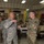 Two military personnel in different camouflage uniforms are engaged in conversation in what appears to be a dining facility or community center. The individual on the left wears the US Air Force ABU (Airman Battle Uniform) pattern, while the individual on the right wears the US Army OCP (Operational Camouflage Pattern) uniform. Multiple other military and civilian personnel are visible seated at tables in the background.