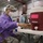 A medical or dental professional wearing military camouflage pants, a purple disposable gown, surgical mask, safety glasses, and a headlamp is seated at a table in what appears to be a gymnasium converted into a temporary medical clinic. Multiple other similarly dressed personnel are visible in the background attending to patients. The setting suggests a military or volunteer medical outreach event.