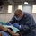 A military dental provider wearing camouflage (OCP) uniform trousers and a blue medical gown is performing a dental procedure on a reclined patient in what appears to be a field or temporary medical clinic setting. The provider is equipped with a headlamp, protective eyewear, surgical mask, and blue nitrile gloves, assisted by a second medical personnel visible on the left. The setting suggests a military humanitarian or readiness dental mission.