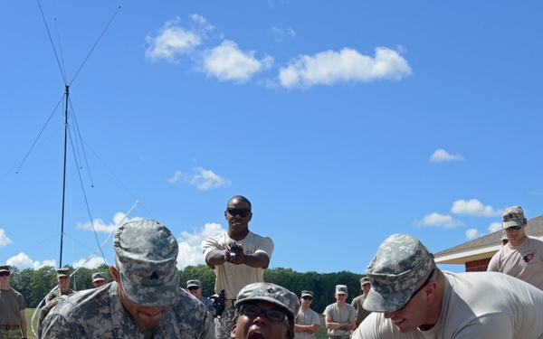 A U.S. Army training exercise is taking place outdoors, showing a soldier being assisted or supported by two other soldiers near blue and black padded mats on the ground. The central soldier appears to be reacting intensely, possibly after completing a physical challenge or receiving a simulated stress inoculation exercise. A group of uniformed and civilian personnel observe in the background.