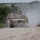 Fighting our nation's wars: one 50lb round at a time A desert-tan painted M1 Abrams main battle tank is moving toward the camera along a gravel road, kicking up a significant dust cloud. A single crew member is visible in the commander's hatch position atop the turret. The setting appears to be a training environment with green vegetation and scrubland on both sides of the road.