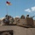 Fighting our nation's wars: one 50lb round at a time The image shows soldiers positioned on top of a desert-tan armored military vehicle, likely an M1 Abrams tank or similar platform. A pennant flag with red and white coloring marked with '3' and 'B' is mounted on the vehicle, indicating a unit designation. Soldiers are equipped with gear, backpacks, and at least one visible rifle, suggesting a field training or operational scenario.