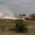 Fighting our nation's wars: one 50lb round at a time A U.S. Army M1 Abrams main battle tank marked with the number '22' is maneuvering across an open grassy training range. To the left, a large explosion with visible flames and a billowing white smoke cloud is visible, consistent with a live-fire training exercise. The scene appears to take place at a military installation, likely in the American Southwest based on the terrain and vegetation.