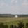 Fighting our nation's wars: one 50lb round at a time Three military tanks are visible on an open grassy firing range, with at least one actively firing its main gun, producing a visible muzzle flash and smoke. The scene appears to be a live-fire training exercise conducted in a flat, open terrain with tree lines in the background. The image was taken from an elevated observation position overlooking the range.