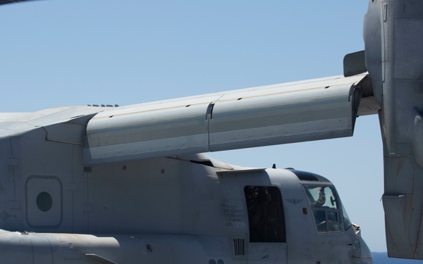 A U.S. Marine Corps MV-22 Osprey tiltrotor aircraft, marked with the number '03', is positioned on the flight deck of a naval vessel at sea. The aircraft is shown in its helicopter mode with nacelles tilted upward, suggesting it is either preparing for takeoff or has recently landed. The image was taken in clear daylight conditions with open ocean visible in the background.