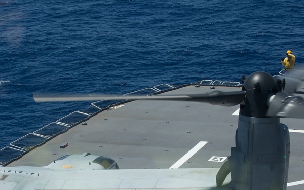 An aerial or elevated view of a military ship's flight deck showing a tiltrotor aircraft, likely a V-22 Osprey, with its rotor assembly prominently visible in the foreground. The flight deck markings, safety netting along the ship's edge, and open ocean are clearly visible. A crew member in a yellow safety vest is visible in the upper right area of the deck.