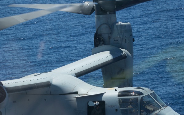 A military tiltrotor aircraft, specifically an MV-22 Osprey, is captured in flight over open ocean water. The aircraft bears the number '03' on its fuselage and is in helicopter mode with rotors tilted upward. At least two crew members are visible through the cockpit and side window.