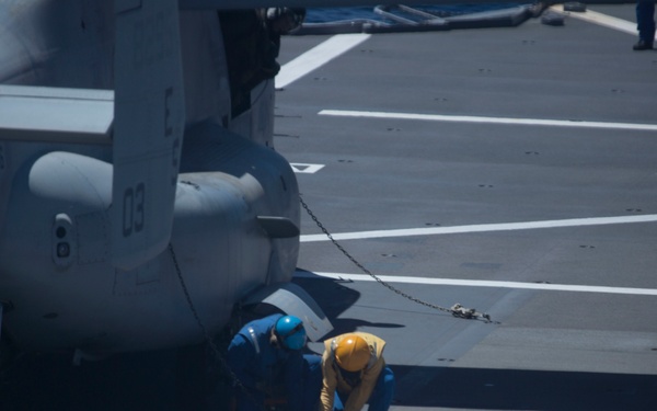 The image shows a naval flight deck with personnel working near a military helicopter, identified by the number '03' on its fuselage. Two deck crew members in colored jerseys and helmets are crouched near the aircraft, likely performing a tie-down or inspection task. A chain securing the aircraft to the deck is clearly visible.