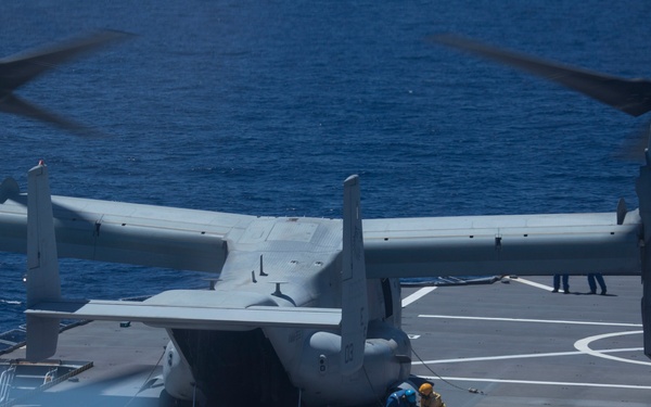 A tiltrotor aircraft, identified as a Bell Boeing V-22 Osprey, is positioned on the flight deck of a naval vessel at sea. The aircraft's rotors are spinning, suggesting it is either preparing for takeoff or has just landed. At least one deck crew member in safety gear is visible near the aircraft performing ground operations.