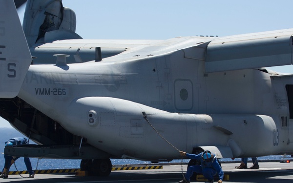 A U.S. Marine Corps MV-22B Osprey tiltrotor aircraft assigned to VMM-266 is parked on the flight deck of a naval vessel at sea. Ground crew members dressed in blue uniforms and helmets are performing tie-down or chaining operations to secure the aircraft to the deck. The aircraft bears the tail code 'ES 03' and bureau number 6295.