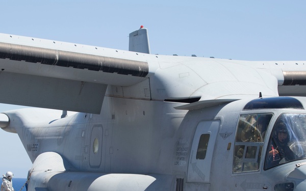 A U.S. Marine Corps MV-22 Osprey tiltrotor aircraft marked '03' is secured on the flight deck of a naval vessel, likely an amphibious assault ship. The aircraft is chained down with tie-down chains and has a pilot or crew member visible in the cockpit. Deck crew in various colored uniforms are visible in the background performing flight deck operations.