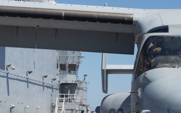 A military aircraft marked '03' is chained down on the flight deck of a naval vessel, likely an aircraft carrier or amphibious assault ship. The aircraft appears to be a C-2 Greyhound or similar carrier-based transport/utility aircraft. Deck crew and support vehicles are visible in the background near the ship's island superstructure.