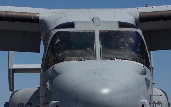 A front-facing view of a U.S. Marine Corps MV-22 Osprey tiltrotor aircraft (numbered '03') chained to the flight deck of a naval vessel. Ground crew members in colored safety jerseys and a fire suppression team in a silver proximity suit are positioned alongside the aircraft. The scene depicts standard flight deck operations at sea under clear sky conditions.