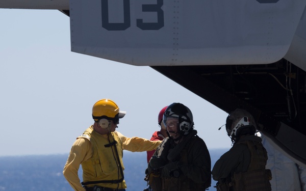 Military personnel are gathered on the flight deck of a naval vessel beneath a tiltrotor aircraft marked with the number '03' and 'VMM' designation, indicating a Marine Medium Tiltrotor Squadron (likely an MV-22 Osprey). A deck crew member in a yellow jersey and yellow helmet appears to be briefing or directing flight crew members wearing flight suits and helmets. The scene takes place at sea under clear, bright conditions.