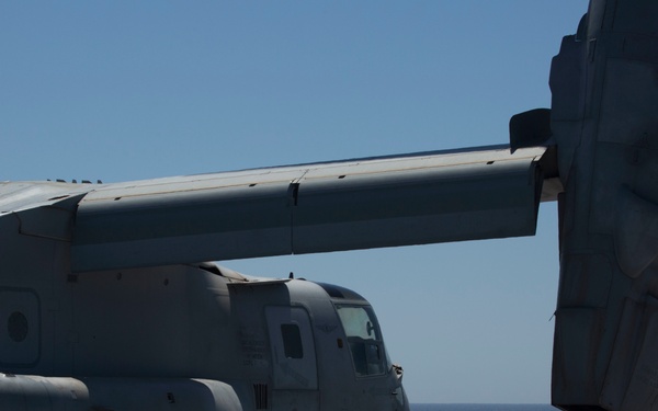 A military tiltrotor aircraft, identified as an MV-22 Osprey, is chained down on the flight deck of a naval vessel at sea. A deck crew member in a yellow jersey and hard hat stands near the edge of the flight deck, apparently observing or signaling a small helicopter visible in the background. The scene takes place under clear blue skies over open ocean.