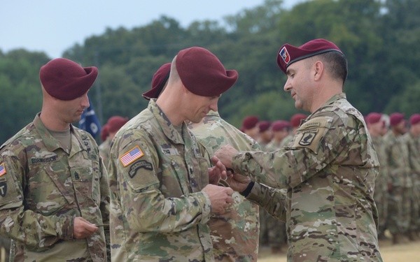 A U.S. Army military ceremony is taking place on a parade field, where a senior officer appears to be presenting an award or decoration to another soldier. All personnel are wearing OCP (Operational Camouflage Pattern) uniforms and maroon berets, indicating they are airborne-qualified paratroopers. A formation of soldiers stands at attention in the background.