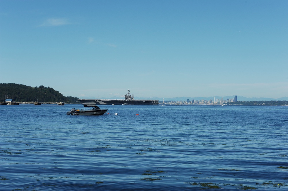 A wide waterway scene showing a large aircraft carrier in the mid-distance, flanked by a forested island on the left and a city skyline on the right. A small motorboat with a bimini top is anchored or idling in the foreground. The image appears to be taken from Puget Sound, with the Seattle skyline visible in the background.