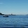 A wide waterway scene showing a large aircraft carrier in the mid-distance, flanked by a forested island on the left and a city skyline on the right. A small motorboat with a bimini top is anchored or idling in the foreground. The image appears to be taken from Puget Sound, with the Seattle skyline visible in the background.
