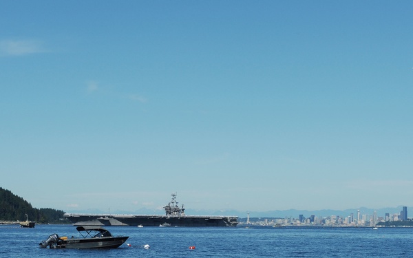 A wide waterway scene showing a large aircraft carrier in the mid-distance, flanked by a forested island on the left and a city skyline on the right. A small motorboat with a bimini top is anchored or idling in the foreground. The image appears to be taken from Puget Sound, with the Seattle skyline visible in the background.