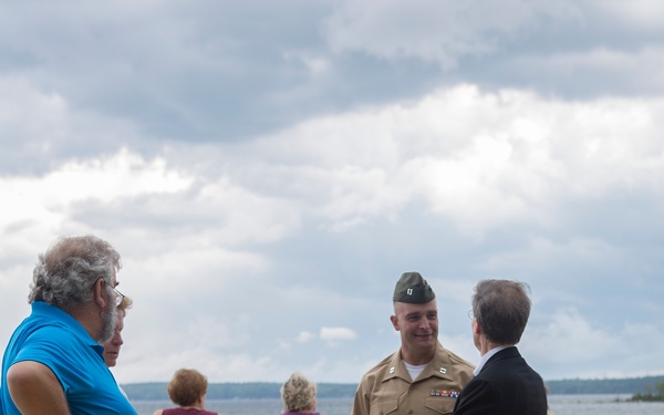 A group of people are gathered on a newly constructed wooden boardwalk or deck overlooking a lake or large body of water. A military officer in tan/khaki uniform with a green garrison cap and service ribbons is engaged in conversation with a man in a dark suit. The scene appears to be an outdoor event or ceremony, with civilians including a child in a Boy Scout uniform also present.