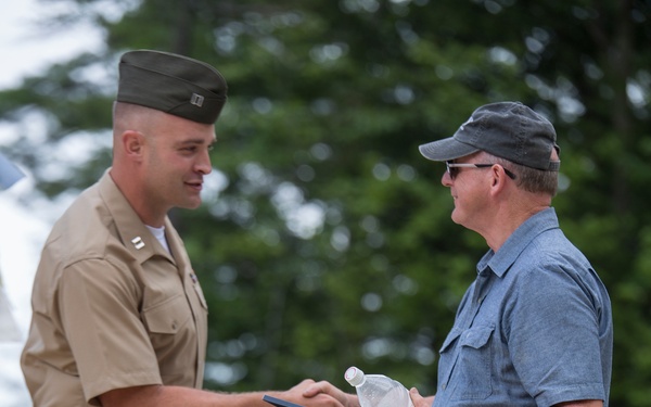 A U.S. Marine officer in service uniform is exchanging or presenting a framed document/plaque to a civilian man outdoors. The Marine is wearing a tan service uniform with a garrison cover and visible rank insignia on his collar. The interaction appears to be a formal presentation or award ceremony taking place on a gravel or sandy surface.