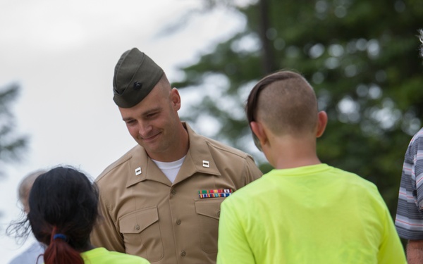 A U.S. Marine officer in service uniform (Charlie/Alpha service uniform) is interacting with two children wearing neon yellow t-shirts, while an elderly civilian man stands nearby holding papers. The setting appears to be an outdoor event among trees, suggesting a community or ceremonial gathering. The Marine is smiling warmly at the children, indicating a positive, friendly interaction.