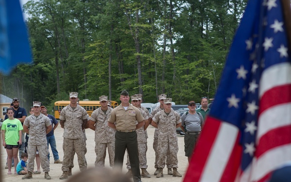 A group of military personnel in camouflage uniforms stand at attention on a sandy outdoor area, facing what appears to be a youth group ceremony. Children in the foreground, appearing to be Cub Scouts or Boy Scouts, are carrying an American flag and other flags during what looks like a flag ceremony or parade. Civilian spectators and a yellow school bus are visible in the background among tall trees.