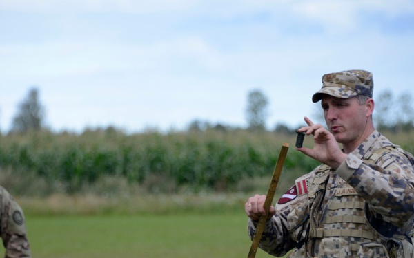 A military instructor in a digital camouflage uniform is conducting an outdoor briefing or training session for a group of soldiers seated on the ground. The instructor is holding what appears to be a wooden stake or training aid while addressing the group in a field setting. The seated soldiers are wearing matching multicam/OCP uniforms with US Army patches visible.