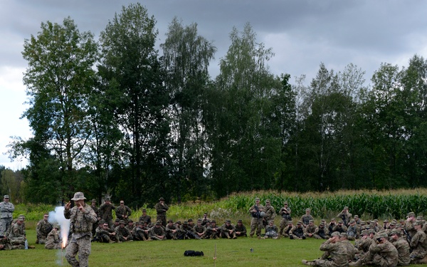 A military training exercise is taking place on an open grass field, with one soldier demonstrating the use of a pyrotechnic device or signal flare while a large group of soldiers observes from a semicircular formation. The setting appears to be a rural training area with trees and what appears to be a cornfield in the background. The overcast sky and green vegetation suggest a temperate climate, possibly in Eastern Europe.