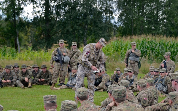 A large group of military personnel in camouflage uniforms are gathered in an outdoor field setting, seated on grass in a semicircle formation. One individual in a distinctive pixelated/digital camouflage pattern stands in the center, appearing to conduct a briefing or demonstration. The setting includes a cornfield and tree line in the background, suggesting a field training exercise.