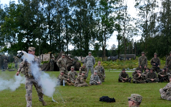 A military training exercise is taking place on an open grass field, where one soldier is demonstrating the use of a smoke or pyrotechnic device to a large group of seated and standing military personnel. The demonstration appears to be an instructional session with multiple service members in various camouflage uniforms observing attentively. A military camp with tents is visible in the background.