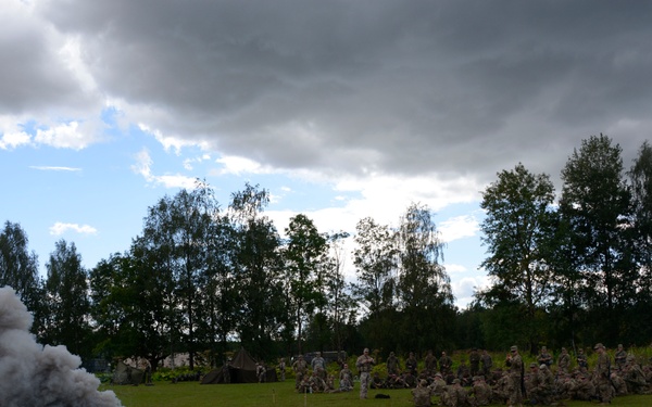 A military training exercise is taking place on an open grass field, with a smoke grenade actively burning and producing a large column of dark gray smoke in the foreground. A large group of soldiers in camouflage uniforms are gathered in the middle distance, some seated and some standing, apparently observing or participating in the demonstration. The setting appears to be a rural training area surrounded by trees under a partly cloudy sky.