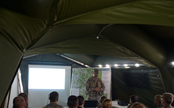 Military personnel are gathered inside a large inflatable field tent for a briefing or operations order. A single presenter stands at the front near a projection screen and a map board, addressing a crowd of uniformed soldiers seated and standing. The setting appears to be a forward operating base or field training exercise environment.
