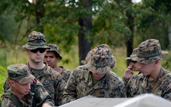 A group of military personnel from at least two nations are gathered around what appears to be a metal panel or boat hull, examining it closely. One soldier is visibly wearing a 'LATVIA' patch on their uniform, while others wear MARPAT (Marine pattern) camouflage consistent with U.S. Marines. The setting is an outdoor wooded or forested area, suggesting a field training exercise or joint military operation.