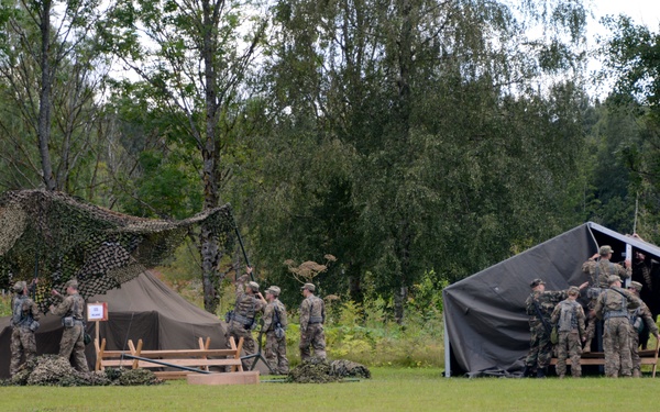 Military personnel in camouflage uniforms are setting up a field camp on an open grassy area bordered by dense trees. Two distinct shelter structures are visible: one with camouflage netting on the left and a larger dark canvas tent/awning on the right. Multiple soldiers are actively working on both structures simultaneously.