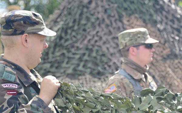 Two military personnel are standing near a camouflage netting structure in an outdoor setting. The soldier in the foreground wears a 'LATVIJA' (Latvia) patch and woodland camouflage uniform with the number 51 visible, while the soldier in the background wears a US Army MultiCam uniform with an American flag patch and sunglasses. This appears to be a joint military exercise or operation between Latvian and US forces.