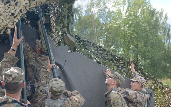 A group of military personnel in mixed camouflage uniforms are collaboratively assembling or erecting a field structure consisting of a black fabric wall panel with camouflage netting overhead. The soldiers appear to represent at least two different nations based on their distinct uniform patterns. The activity takes place in an outdoor field environment with trees visible in the background, suggesting a training exercise or field operation setup.