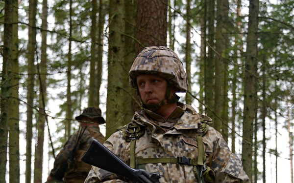 Two soldiers in digital camouflage uniforms are conducting a patrol or tactical movement through a dense coniferous forest. The foreground soldier is equipped with a rifle, helmet, and tactical gear, and appears alert and scanning the surroundings. A second soldier is visible in the background, similarly equipped.