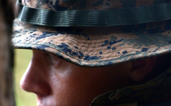 A close-up profile shot of a military personnel wearing MARPAT (Marine Pattern) digital camouflage boonie hat and gear. The subject appears to be in a field or outdoor training environment, partially obscured behind cover near a tree. The image captures a focused, alert expression suggesting a tactical or training scenario.