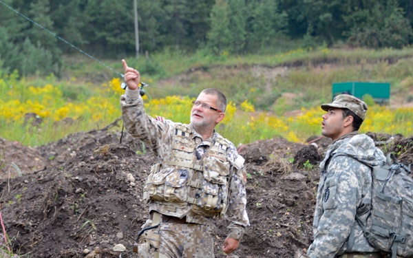 Two military personnel in different camouflage uniforms are standing in what appears to be a field training area or exercise ground. The soldier on the left, wearing a pixelated/digital camouflage pattern with a tactical vest, is pointing upward while appearing to brief or instruct the other soldier. The second soldier, wearing US Army ACU (Army Combat Uniform) pattern with a backpack, is observing the direction being indicated.