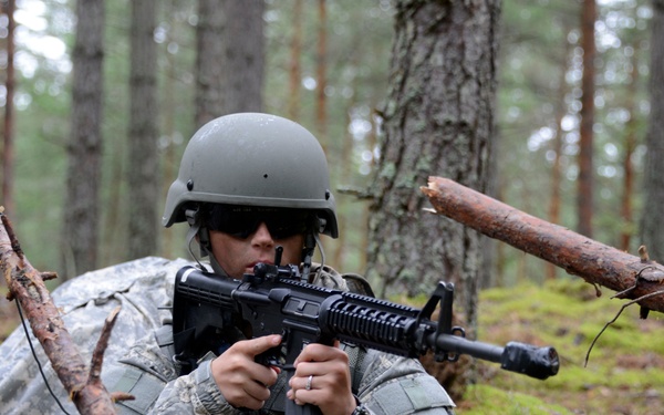 A soldier in U.S. Army ACU (Army Combat Uniform) pattern camouflage is positioned low to the ground in a forested area, holding an M4/M16-style carbine in a ready position. The soldier is wearing a standard ACH (Advanced Combat Helmet) in olive drab and protective eyewear. The setting appears to be a woodland training exercise based on the environment and posture.